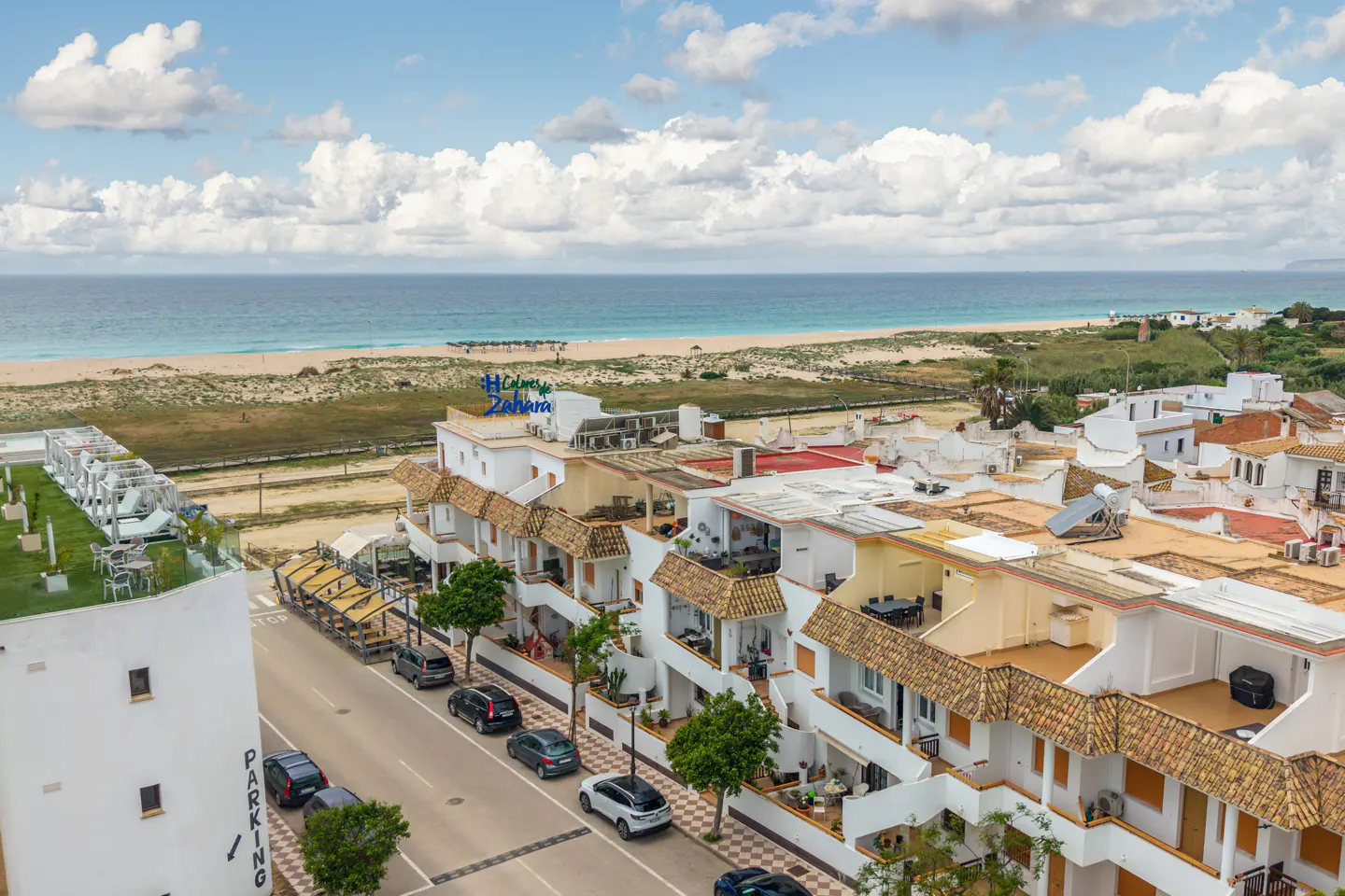 Coastal view of Zahara, Spain, featuring white buildings with terracotta roofs, a sandy beach, and the blue Atlantic Ocean under a cloudy sky.