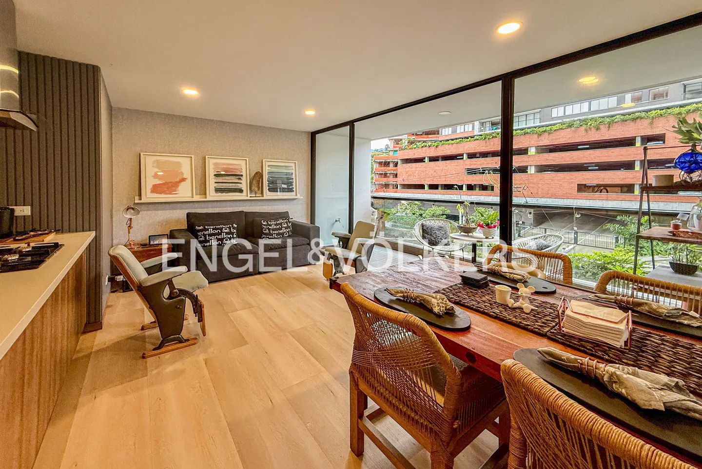 Modern apartment with a wooden dining table, grey sofa, and floor-to-ceiling windows overlooking a brick building.