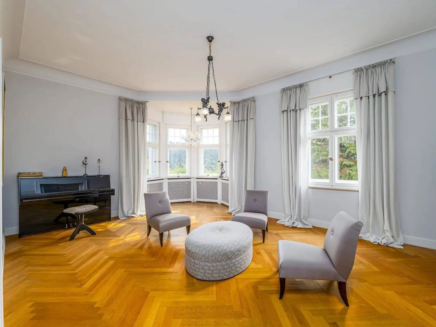 A bright living room with a piano, three chairs, an ottoman, and large windows.