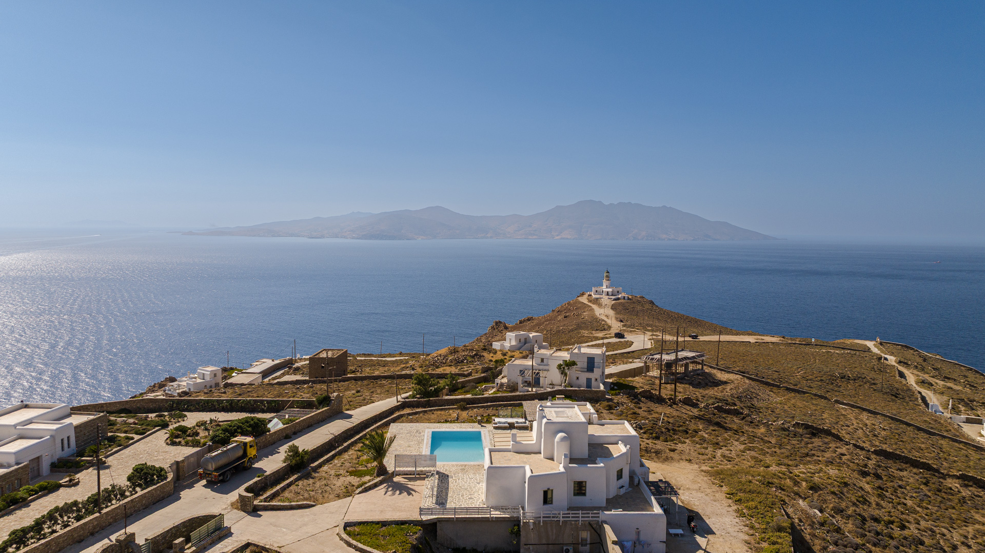 Aerial view of a white villa with a pool on a Greek hillside, overlooking the blue Aegean Sea and a distant lighthouse.