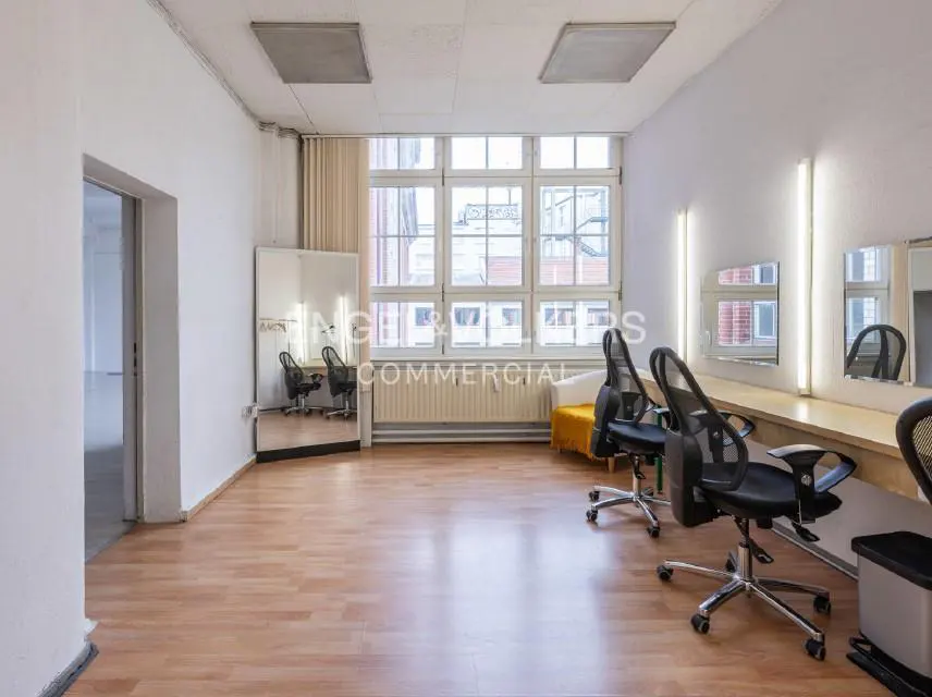 Bright, empty studio with wood floors, white walls, and large windows. Black chairs face mirrors with lights above. A yellow bench sits near the window.