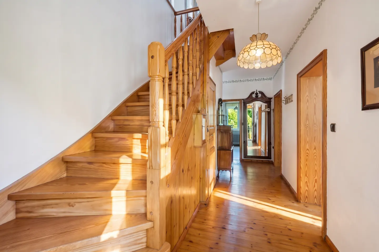 A wooden staircase with a banister leads up from a hallway with light wood floors, a pendant lamp, and a hallway mirror.