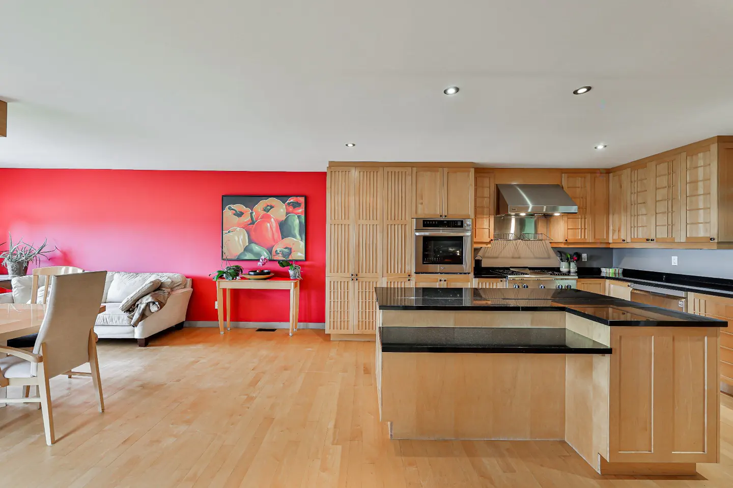 A bright, open-concept kitchen with light wood cabinets, black countertops, and stainless steel appliances. A red accent wall features a colorful painting.
