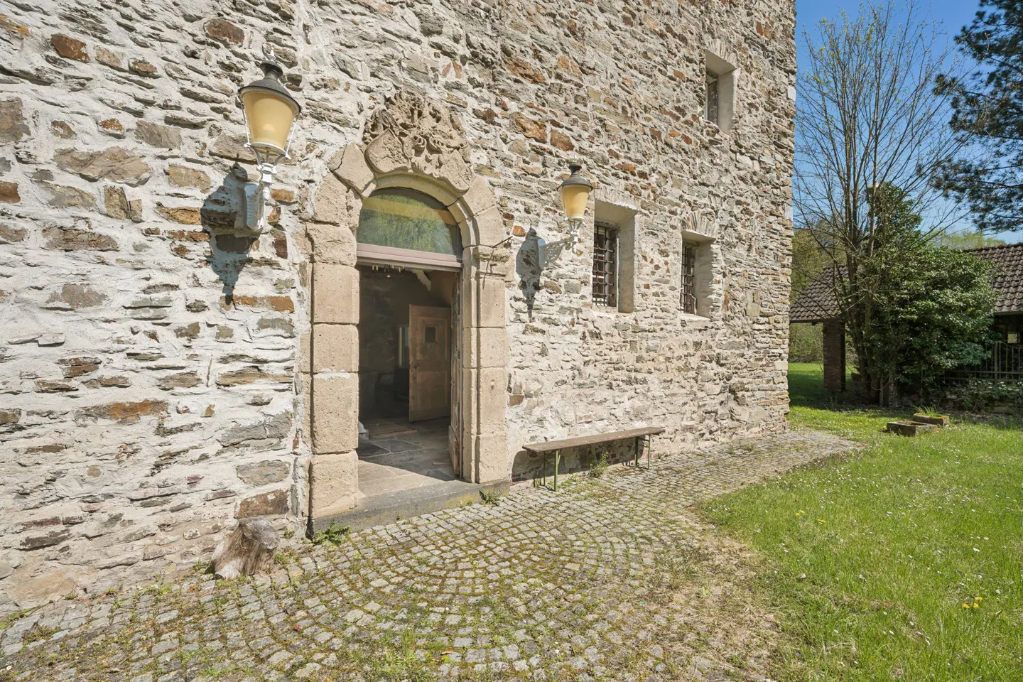 Stone building exterior with arched doorway, flanked by lamps. Cobblestone path leads to the entrance, with a bench nearby. Green lawn and trees in the background.