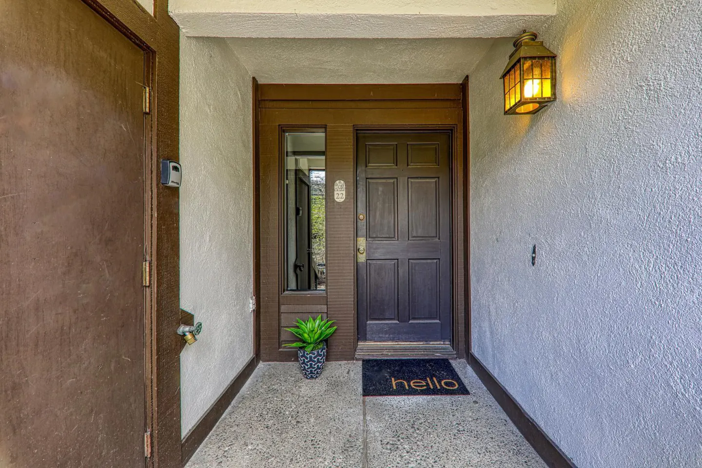 A brown front door with a "hello" mat, a potted plant, and an outdoor light fixture.
