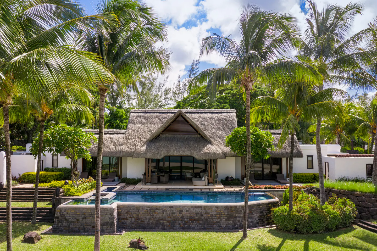 Luxury villa with a thatched roof and a blue pool, surrounded by palm trees and green grass.