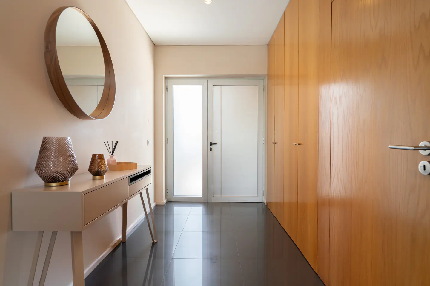 Hallway with a console table, round mirror, and wooden cabinets. The floor is dark gray tile, and a white door is at the end of the hall.