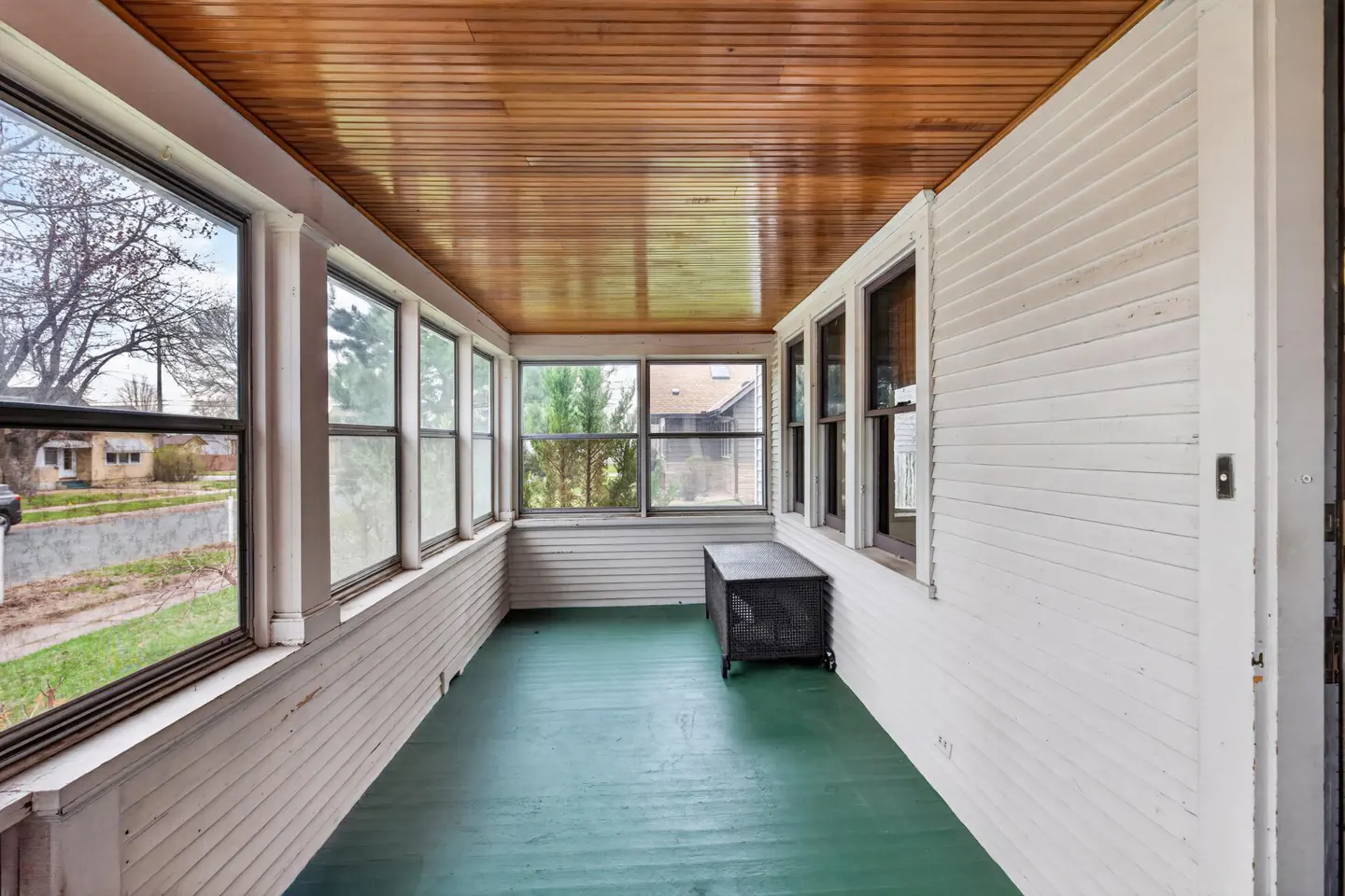 Enclosed porch with green floor, white walls, and wood ceiling. Windows show neighborhood views. A black metal bench sits against the wall.