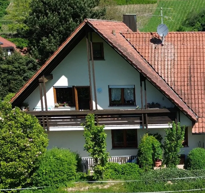 A two-story white house with a red tile roof and a balcony surrounded by green bushes.