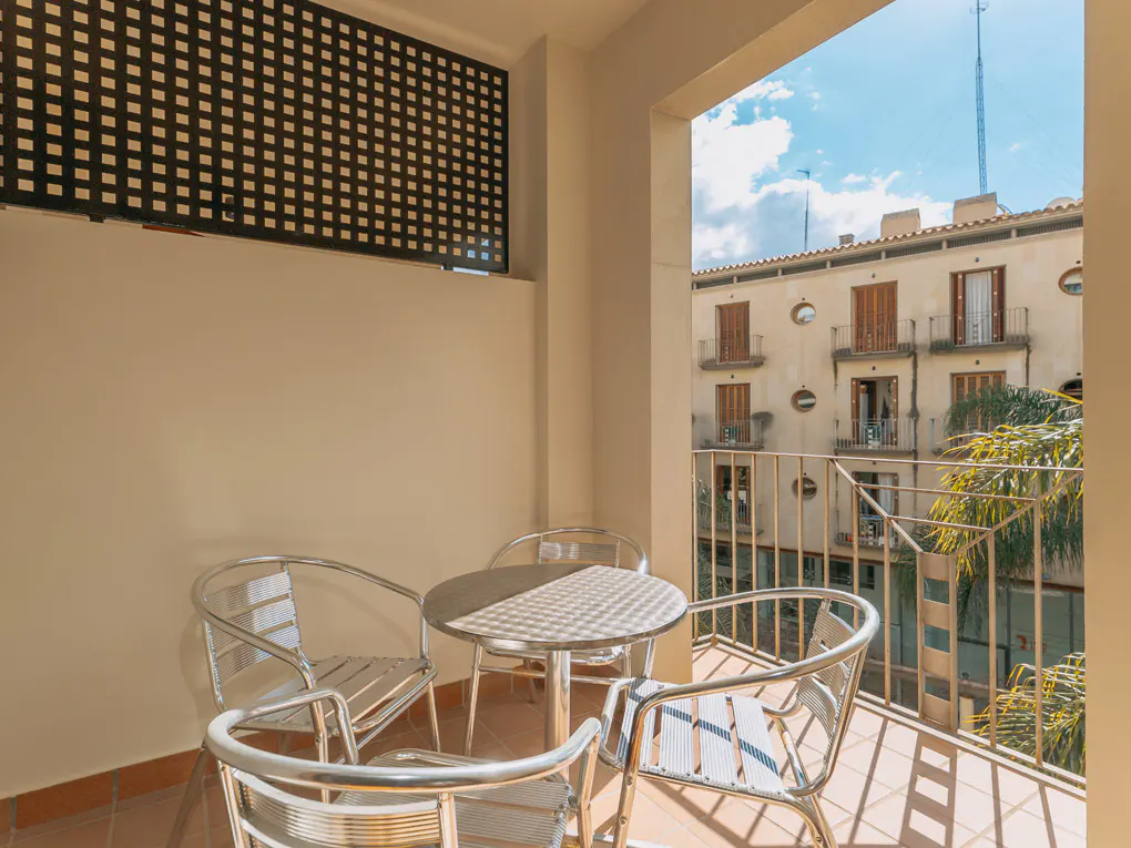 Balcony with a round table and four chairs. A building is visible in the background with blue sky and palm trees.
