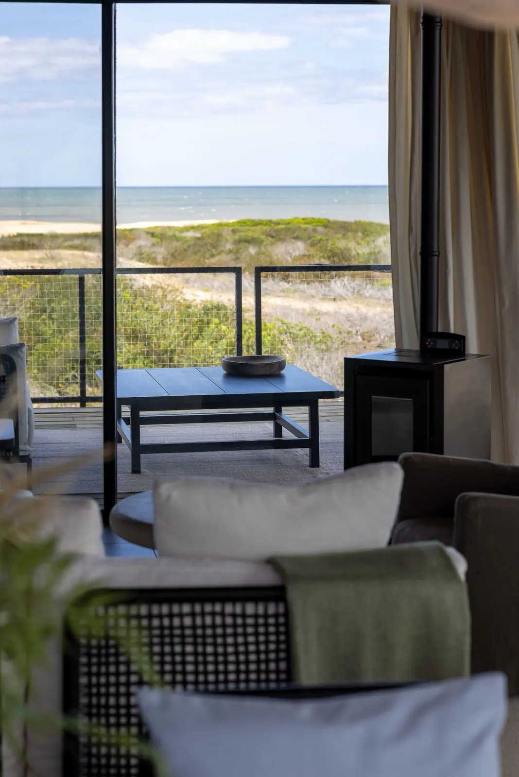 Living room with a black table and fireplace, overlooking a sandy beach and ocean through a large window. Neutral tones and natural light.