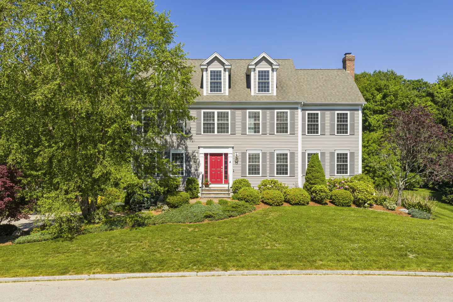Two-story gray colonial home with a bright red front door, dormer windows, and a lush green lawn under a clear blue sky.