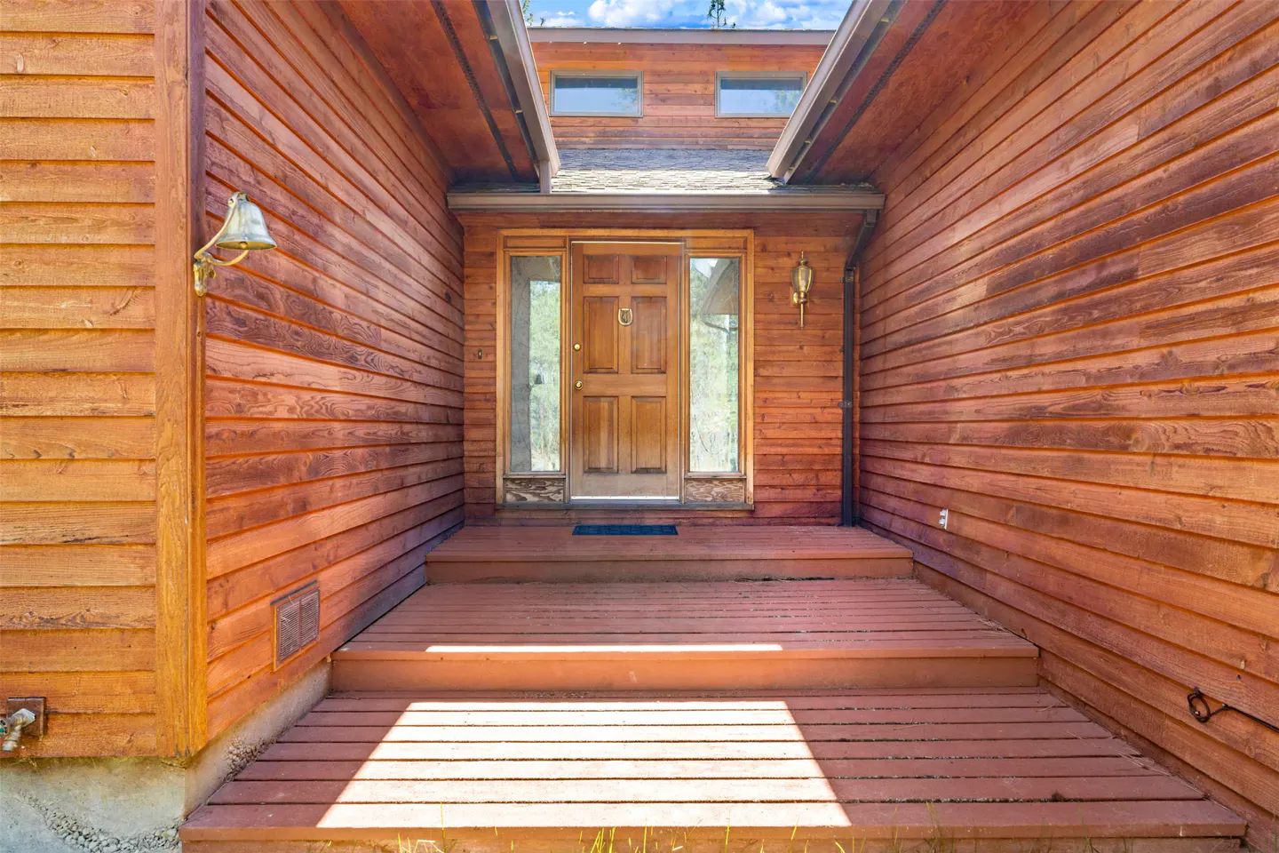 Exterior of a wood-sided house with a wooden door, sidelights, and a wooden porch with steps. A brass bell is on the left.