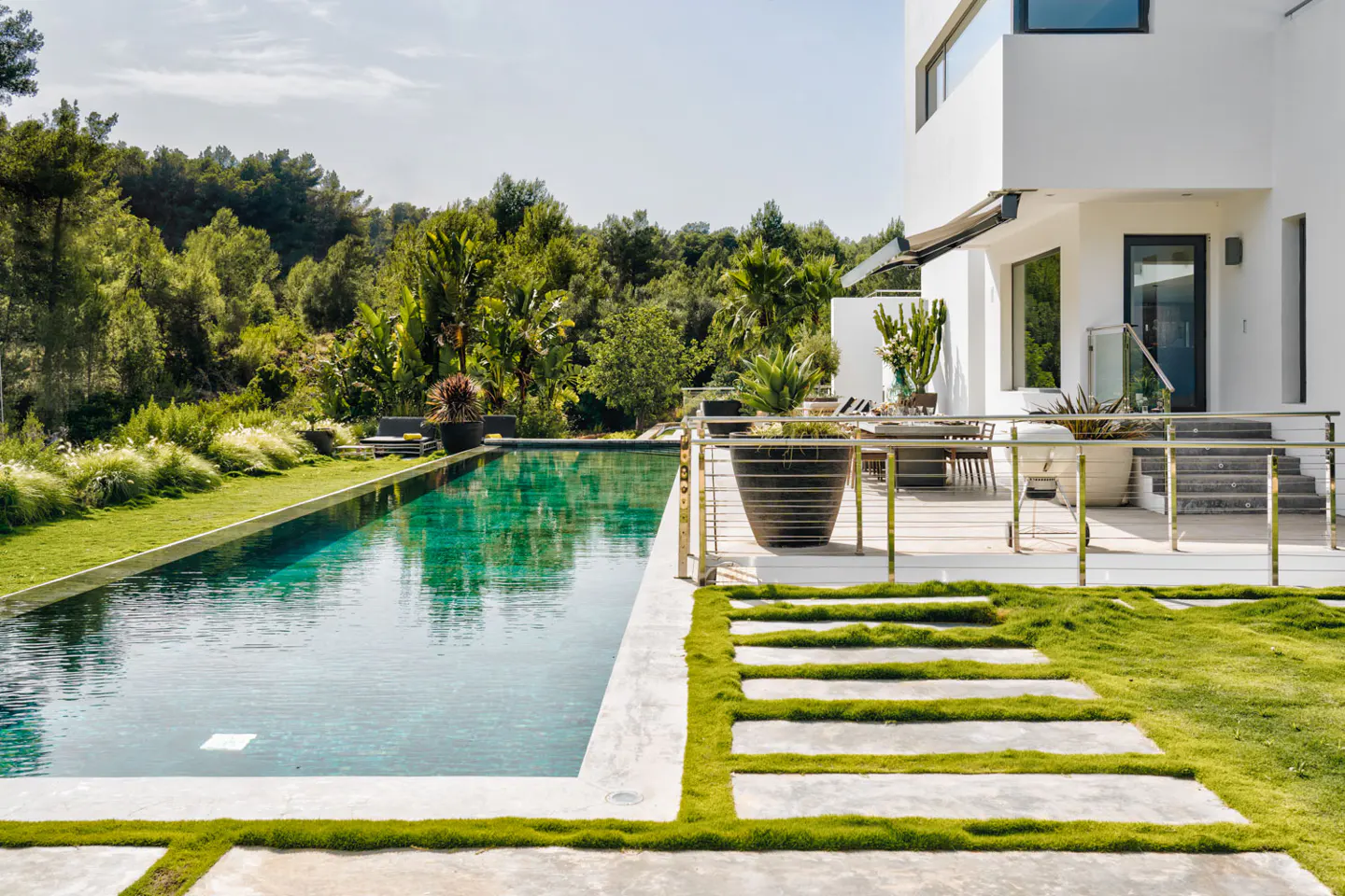 A modern white house with a pool and lush greenery. Stone steps lead to a patio with plants and outdoor furniture.