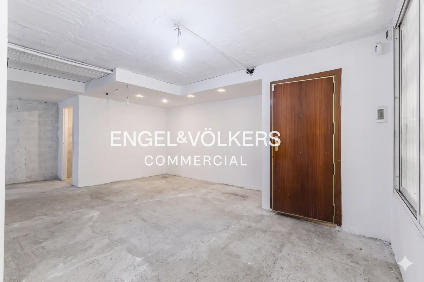 Empty commercial space with white walls, concrete floor, and a brown door. Engel & Völkers logo is superimposed on the image.