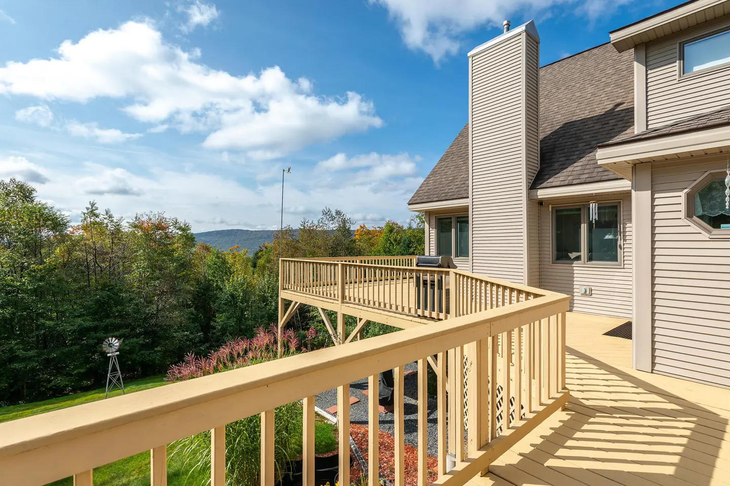 Tan house with a wooden deck overlooking a green lawn, trees, and distant mountains under a blue sky with white clouds.