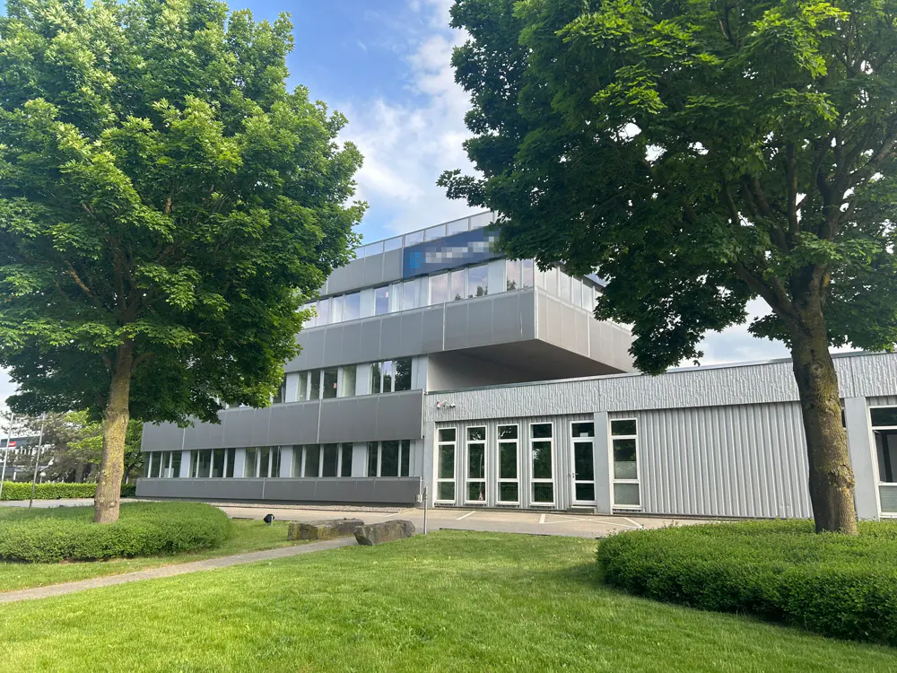 A modern, gray office building with large windows, framed by green trees and a lawn.