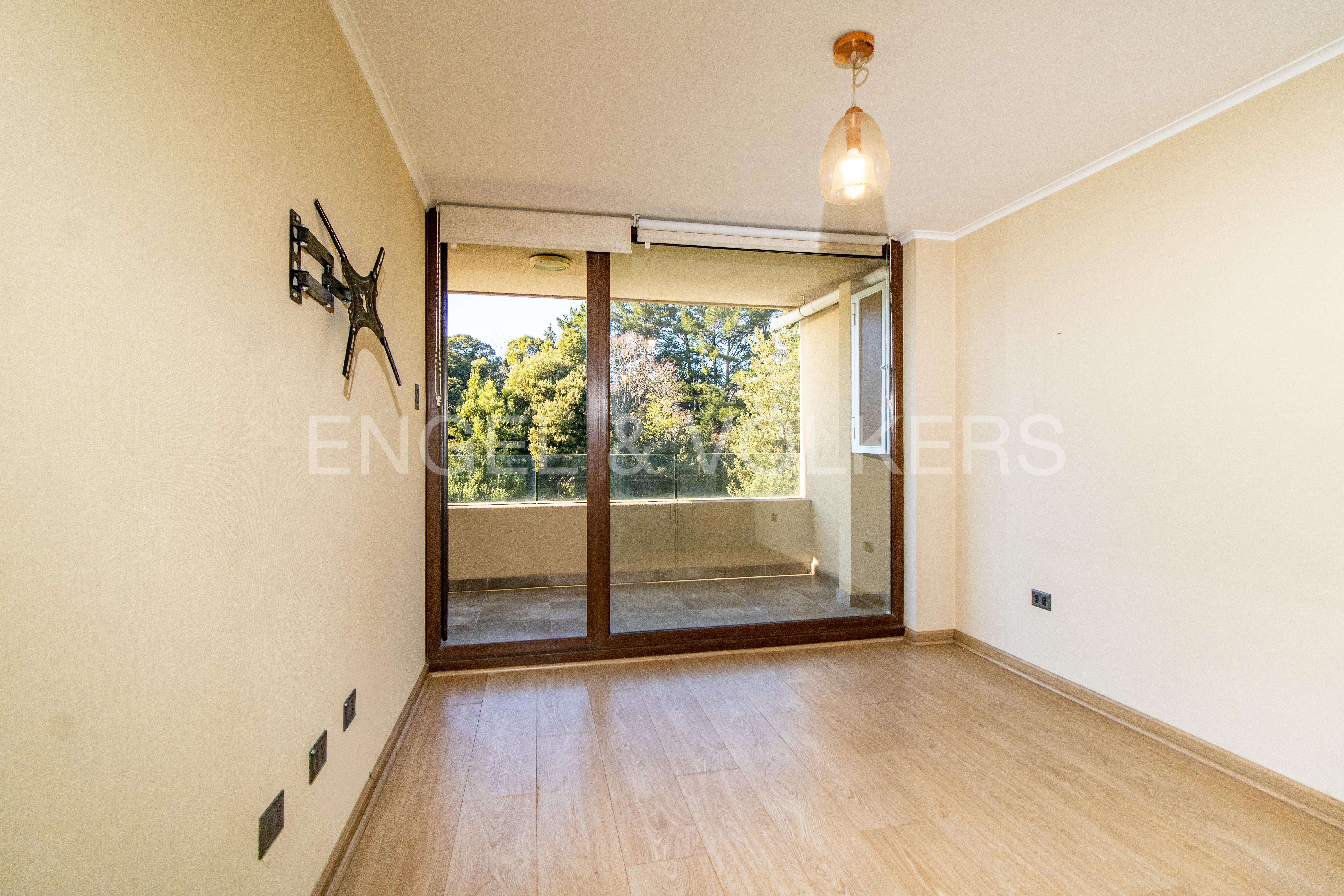 Empty room with light wood floors, beige walls, a wall-mounted TV bracket, and a glass door leading to a balcony with trees.