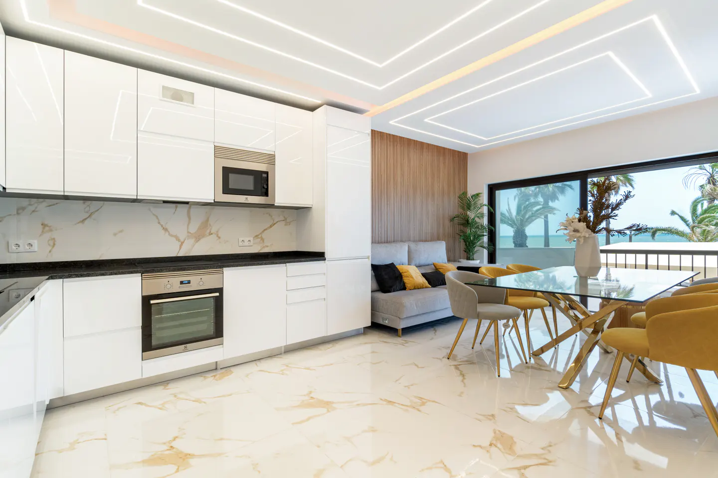 Modern kitchen with white cabinets, marble floors, and a dining area with yellow chairs overlooking a palm tree view.