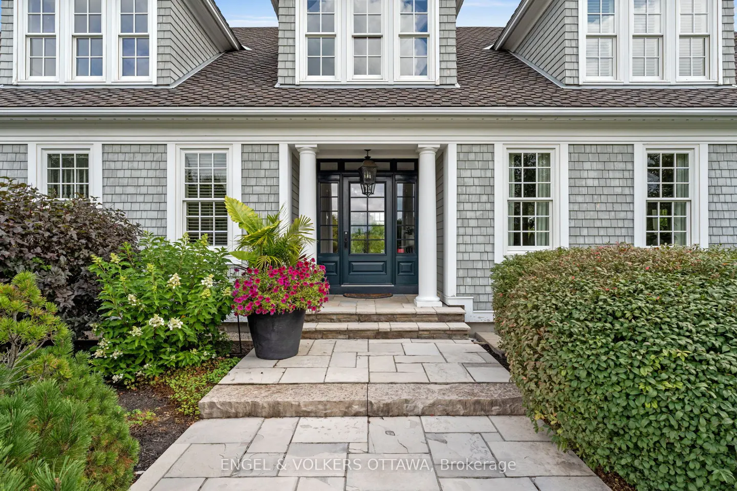 Front entrance of a gray shingle house with white trim, columns, and a dark blue door. Stone walkway with flower pot.