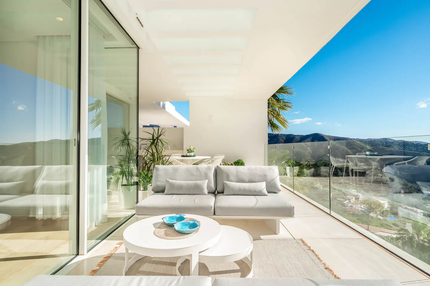 A bright, modern patio with white furniture, a glass railing, and a scenic mountain view.