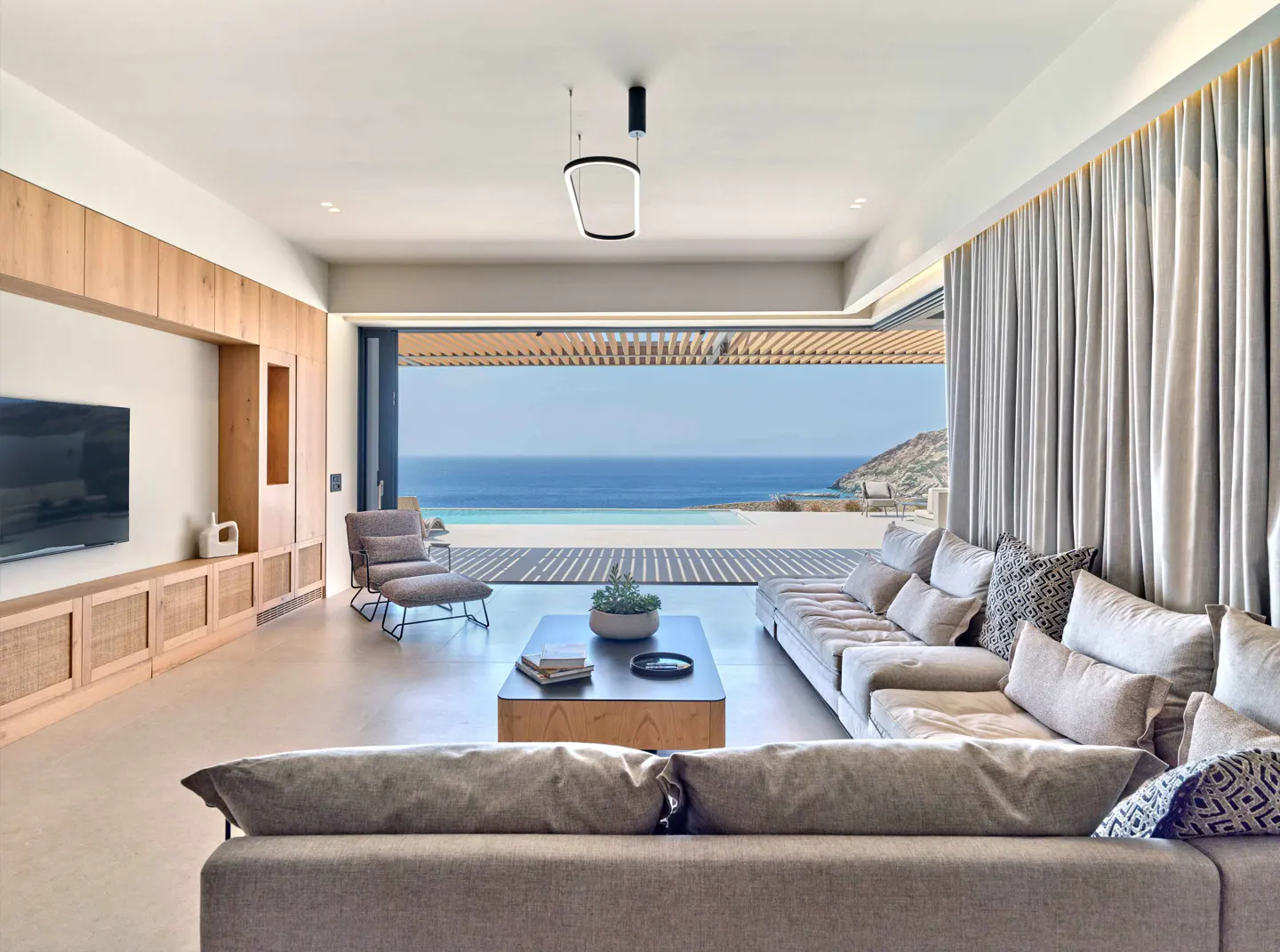 Living room with gray sofas, wood cabinets, and ocean view. Sliding glass doors open to a patio with a pool and blue sea.