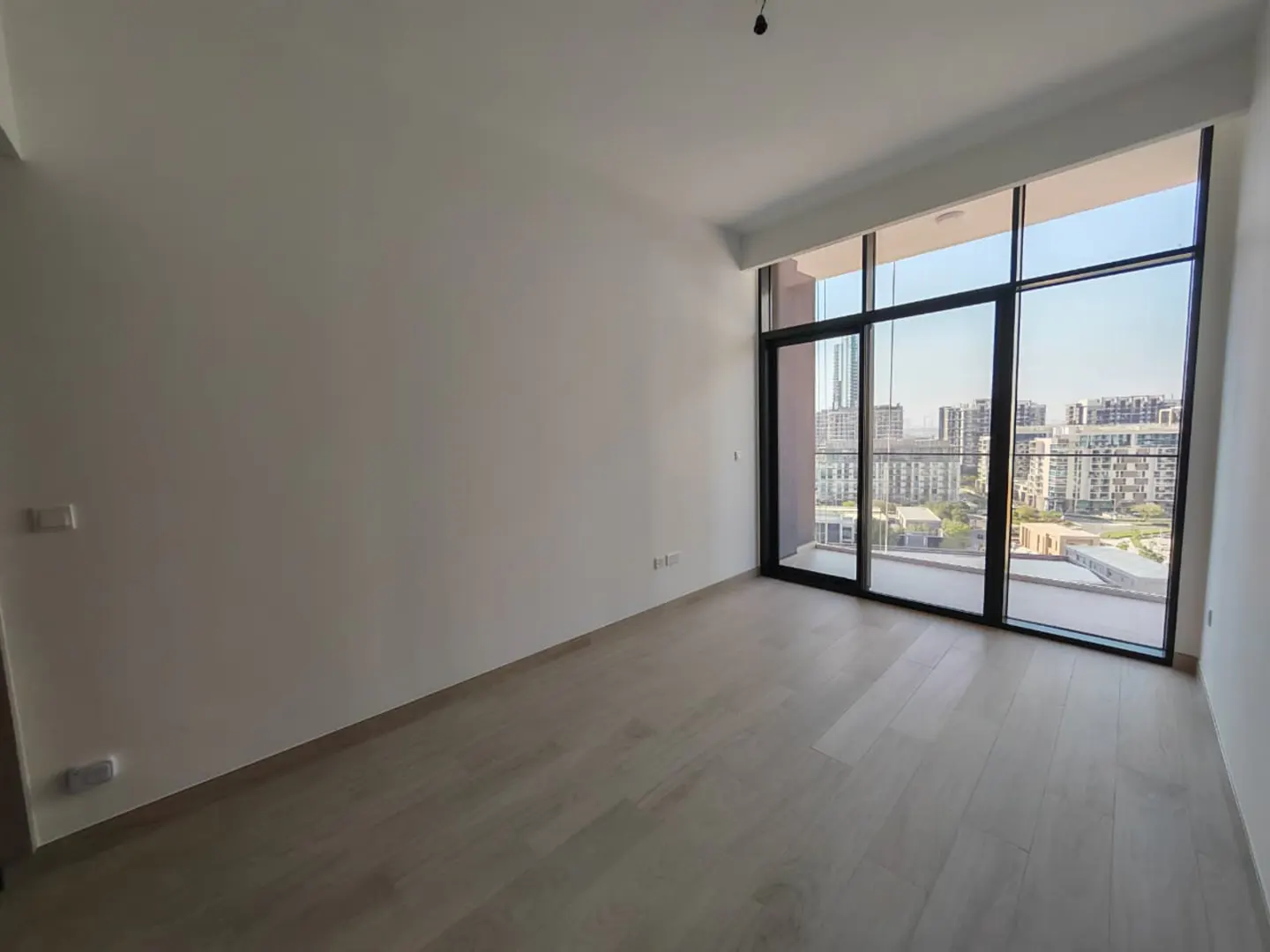 Bright, empty room with light wood floors and white walls. Large black-framed windows offer a city view.