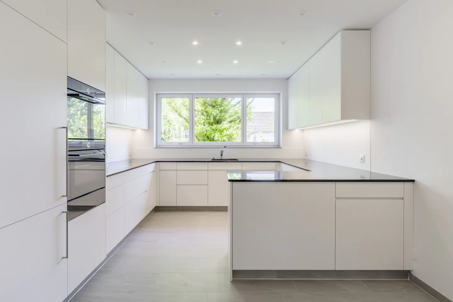 Modern U-shaped kitchen with white cabinets, black countertops, built-in oven, and a window overlooking green trees.