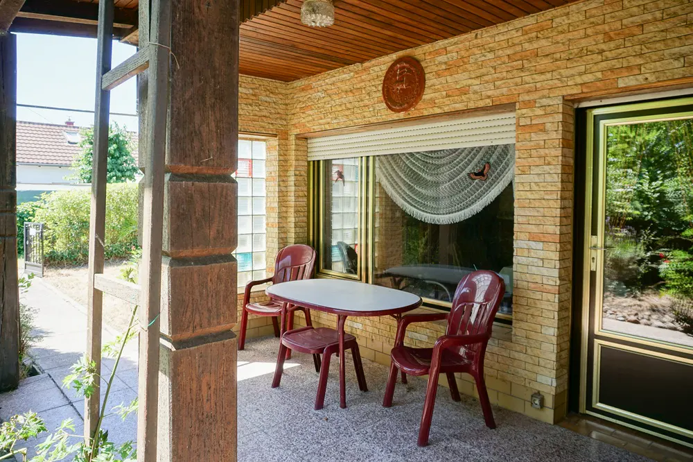 Covered porch with brick walls, wood ceiling, and stone floor. A white table and three red plastic chairs sit near a window with a lace curtain.