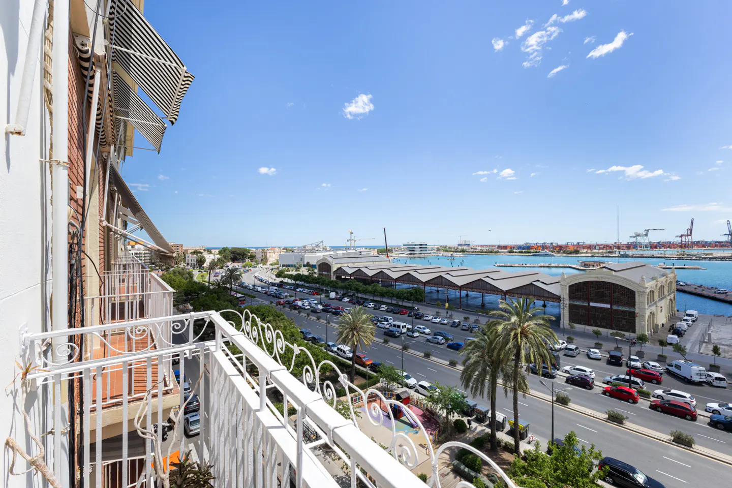 View from a white balcony overlooking a busy street, palm trees, and the Valencia port with blue water.