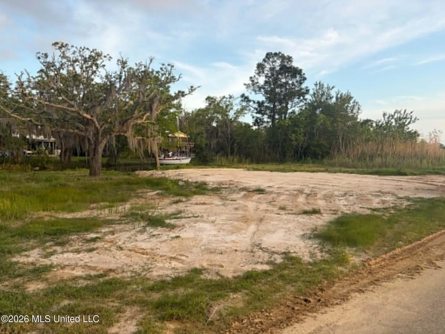 Vacant lot with sandy soil, tire tracks, and sparse grass. Trees and a boat are visible in the background.