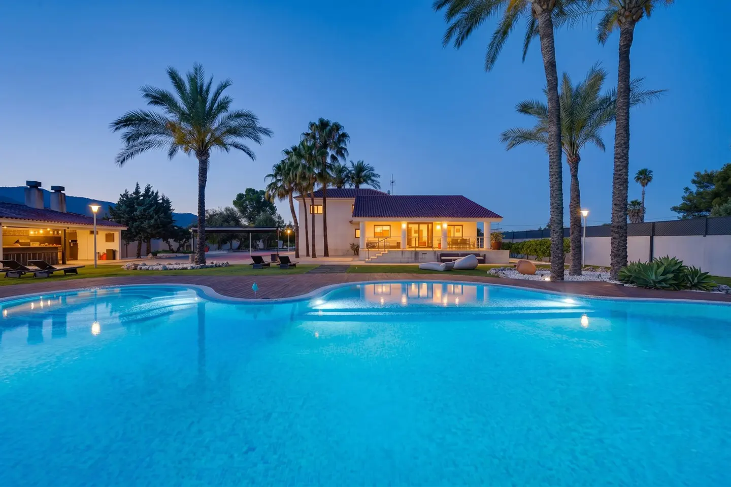 A turquoise pool reflects lights and a house with a red roof at dusk, surrounded by palm trees.