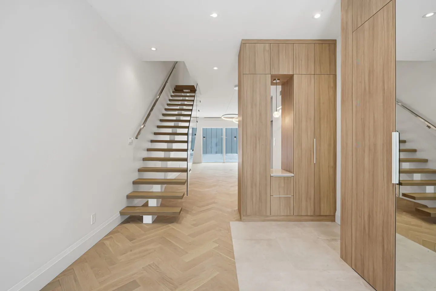 Bright entryway with wood herringbone floors, white walls, and a floating staircase with glass railing. A built-in wood cabinet is on the right.