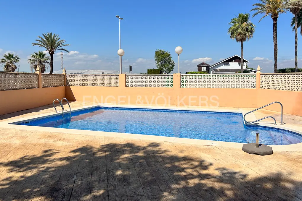 A blue tiled swimming pool with metal ladders, surrounded by a peach-colored wall and palm trees under a clear blue sky.