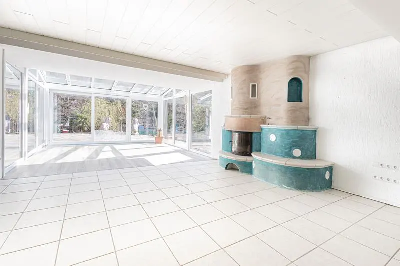 Bright sunroom with white tiled floors, a unique teal and beige fireplace, and large glass walls overlooking a garden.
