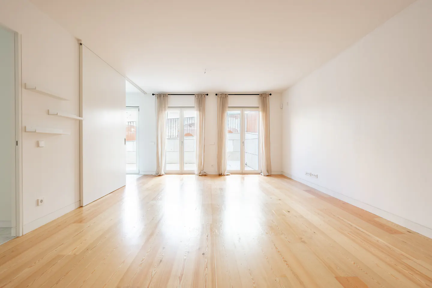 Bright, empty room with light wood floors, white walls, and two sets of French doors with beige curtains.
