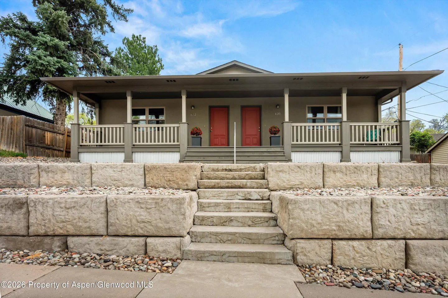 Tan duplex with red doors, a covered porch, and stone retaining walls under a blue sky.