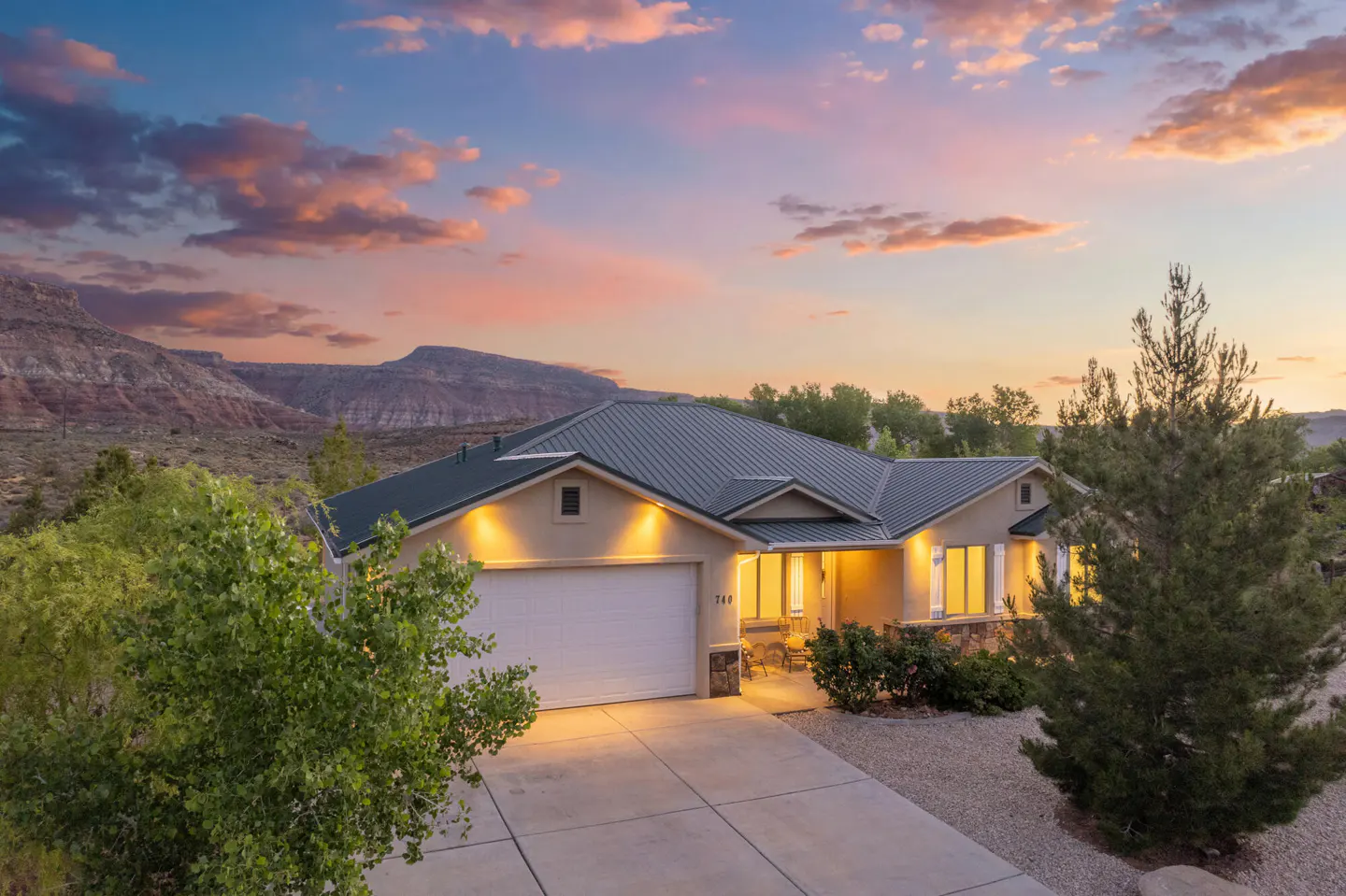 Tan single-story home with a green roof, white garage door, and desert landscape at sunset. Mountains are visible in the background.