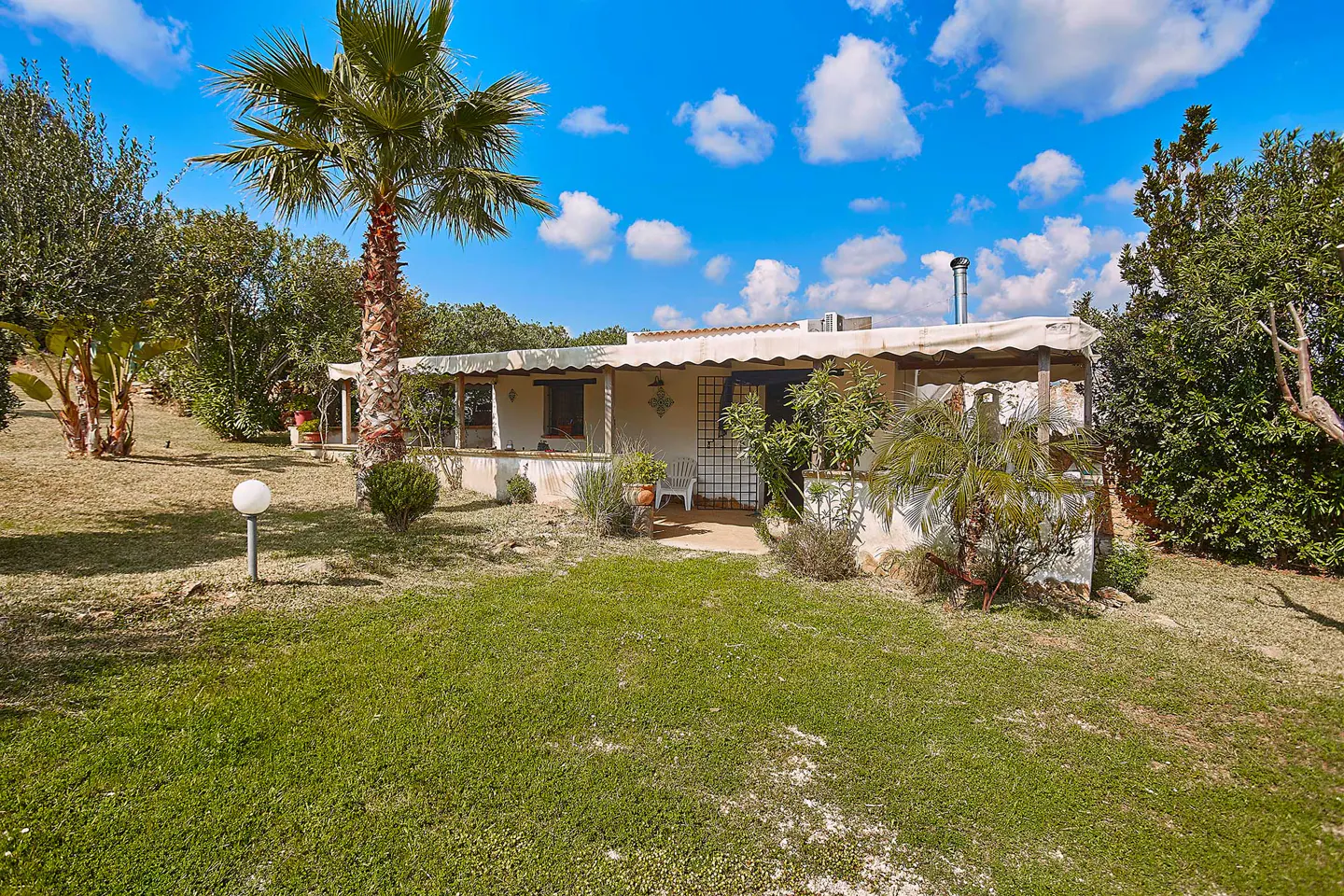 A white, single-story house with a covered porch, surrounded by green lawn, trees, and a tall palm tree under a blue, cloudy sky.
