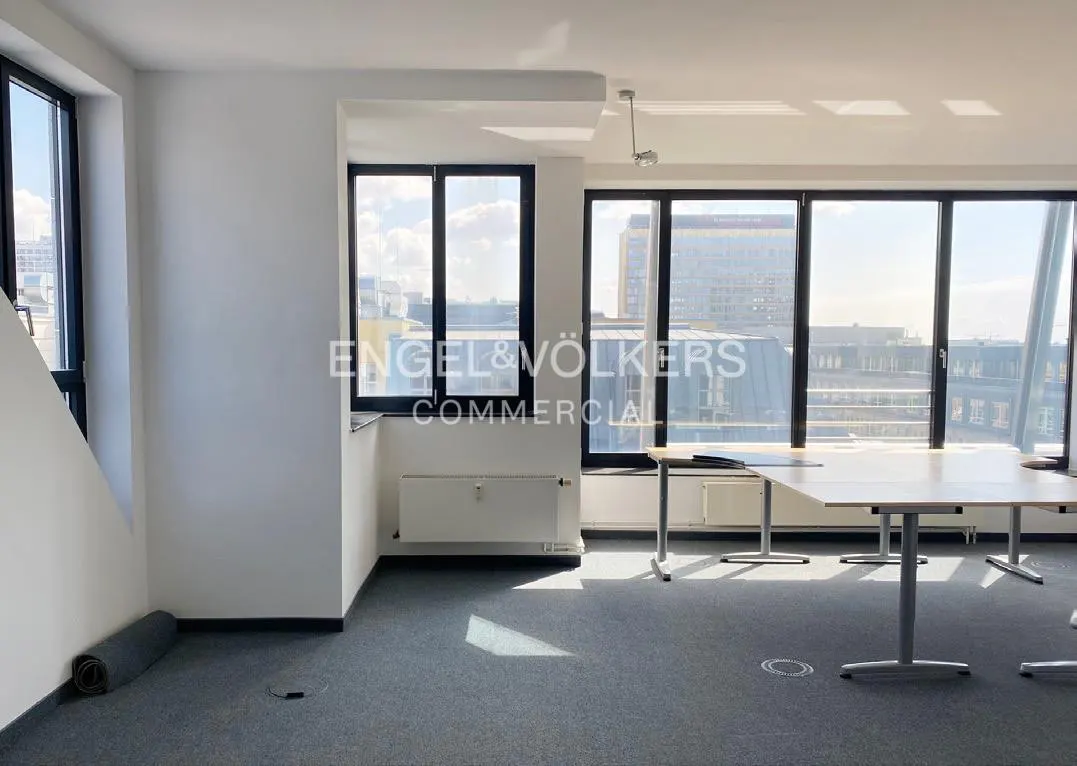 Bright office space with white walls, gray carpet, and large windows overlooking a city. Tables are set up for work. A rolled-up carpet sits in the corner.