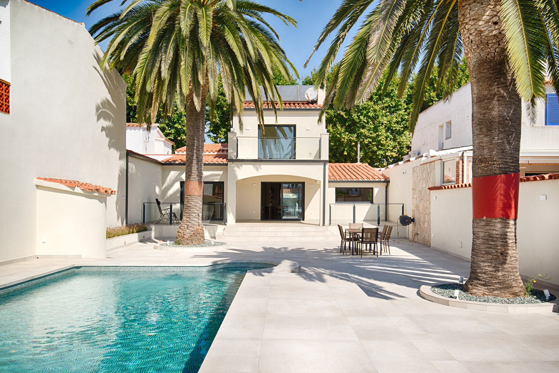 Sunny courtyard with a swimming pool, palm trees, and a modern two-story villa with a patio dining set.