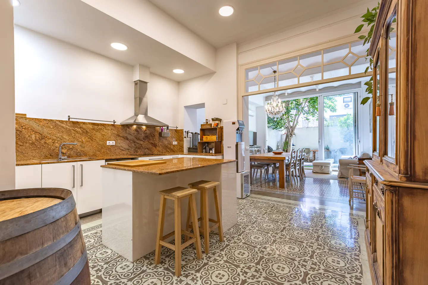 A bright, open kitchen with white cabinets, granite countertops, and a center island with two wooden stools. A dining area is visible through an open doorway.