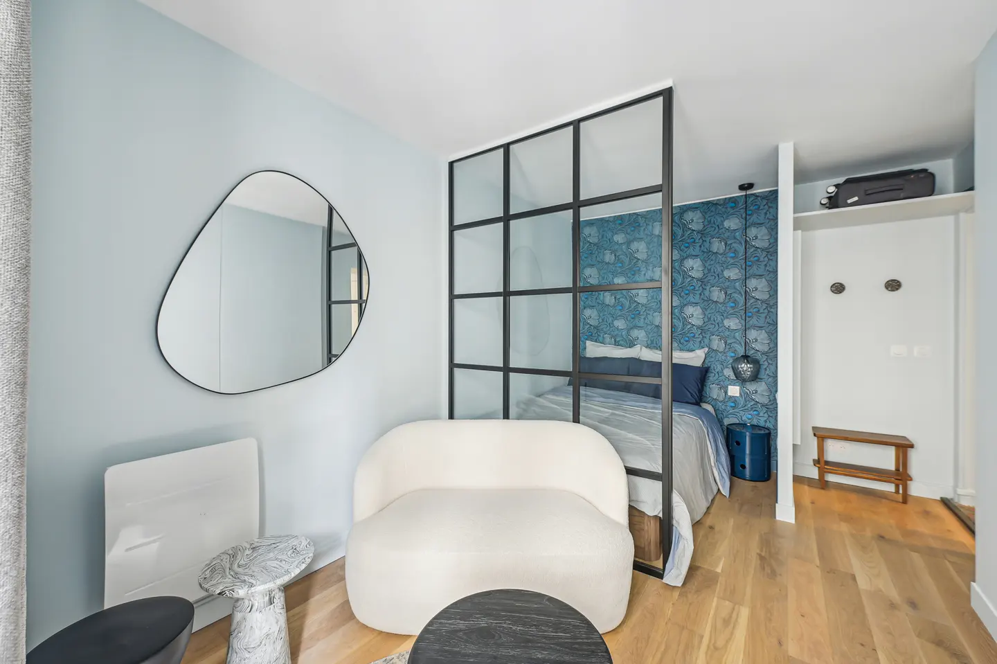 A modern studio apartment with a white chair, black framed glass divider, and blue floral wallpaper.