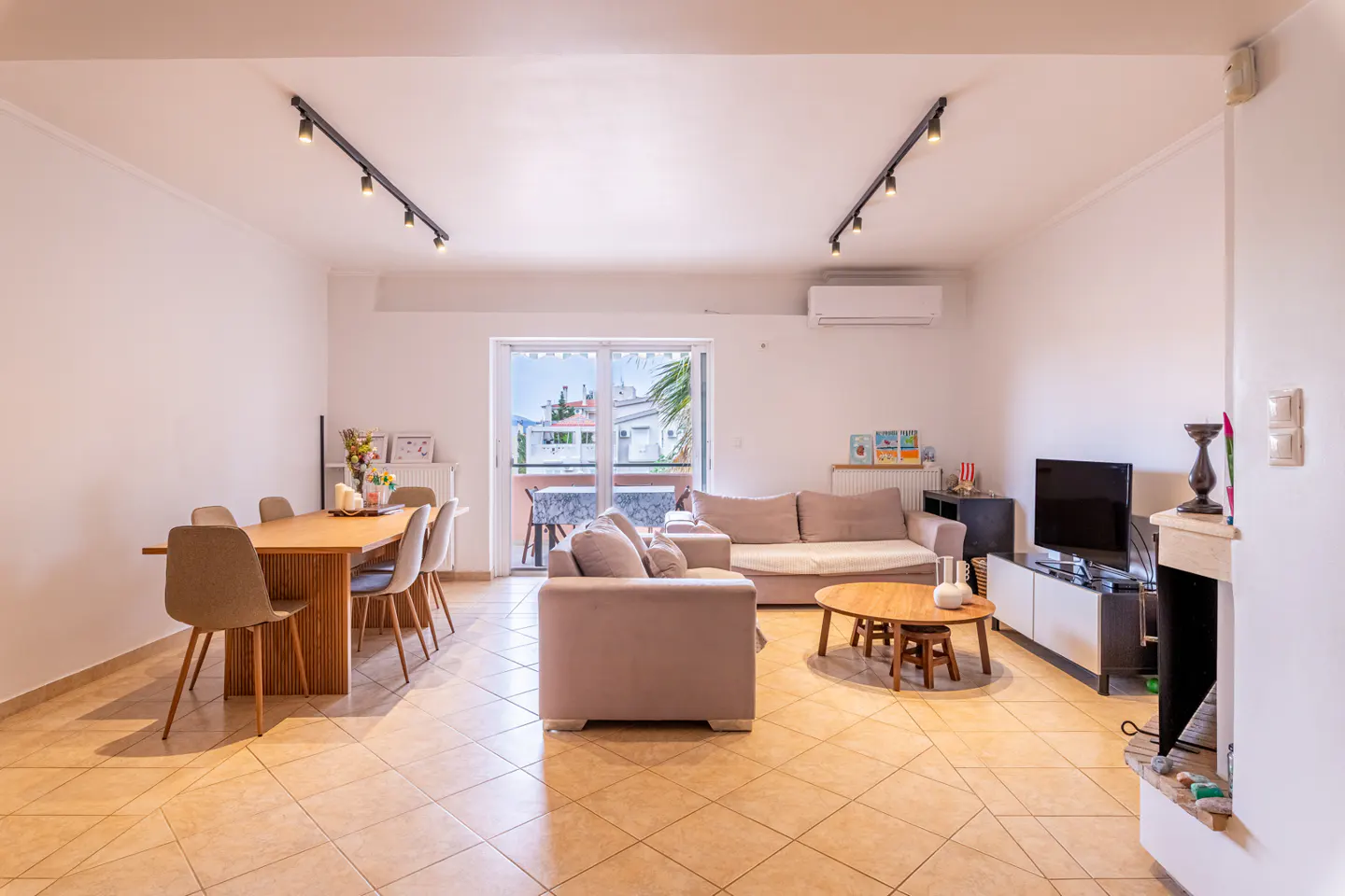Bright open-plan living and dining room with beige tile floors, a light sofa, wooden dining table, and balcony access.