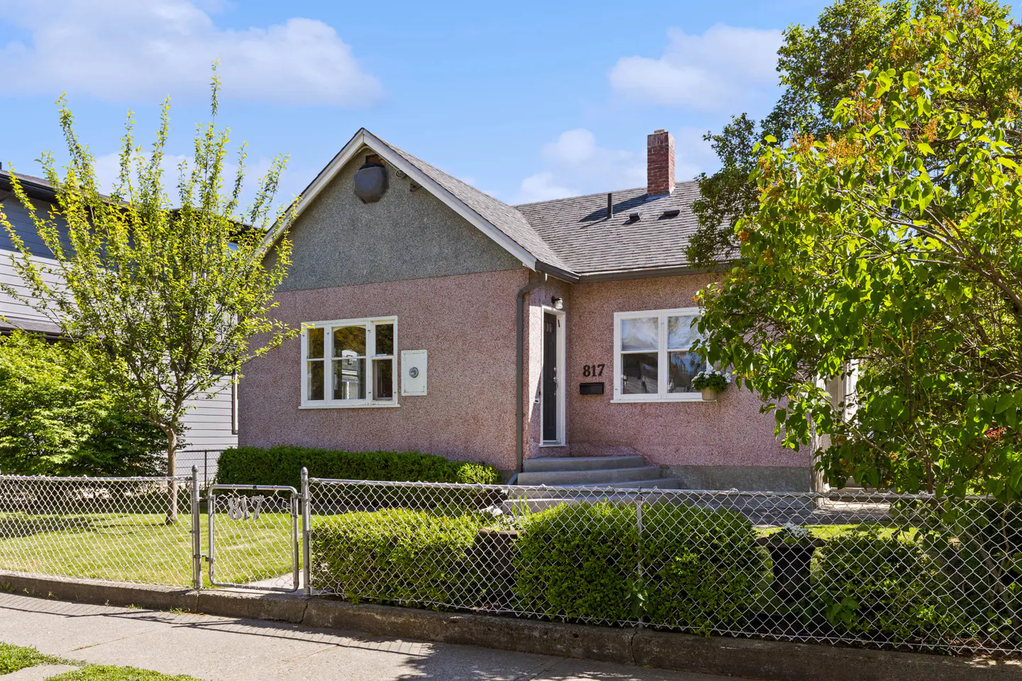 A pink stucco house with white trim, a gray roof, and a brick chimney, surrounded by green trees and a chain-link fence.