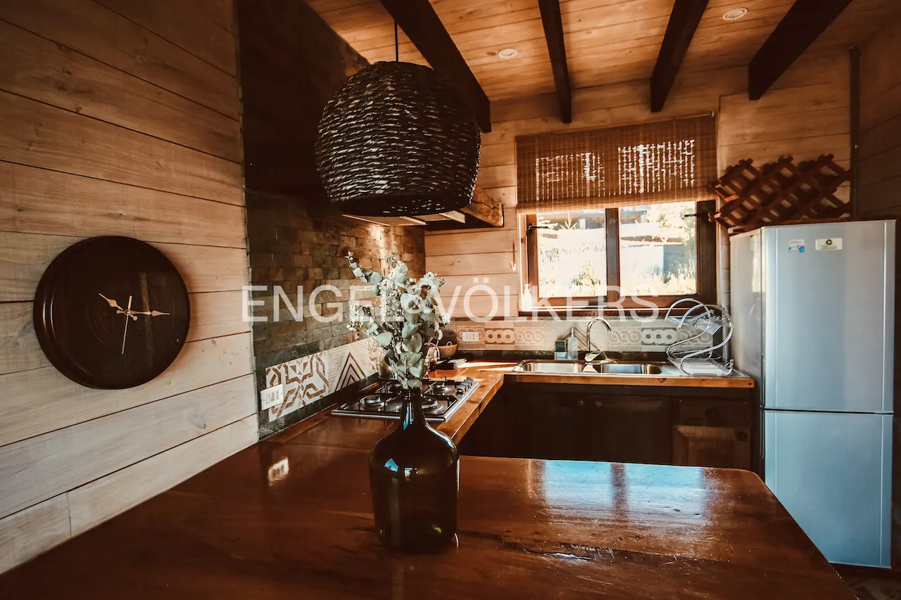 A rustic kitchen with wood walls, a dark wicker light fixture, and a wooden countertop. A black clock hangs on the wall.