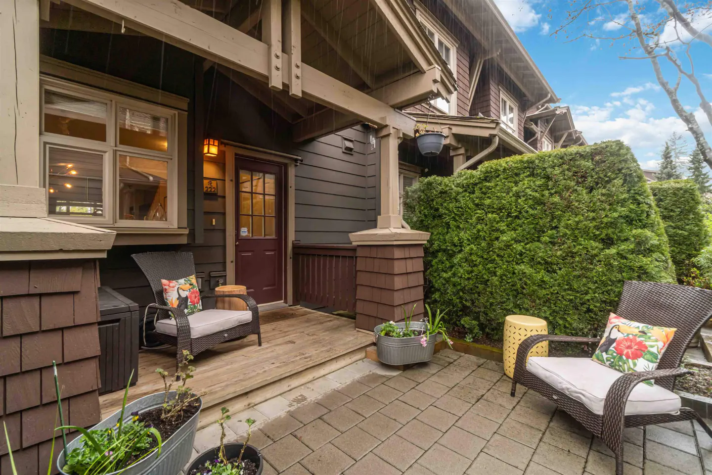 Exterior view of a brown house with a porch featuring two chairs with floral pillows and potted plants.