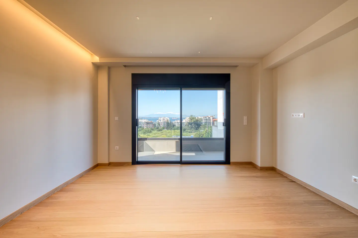Empty room with light wood floors, beige walls, and a large sliding glass door with a view of a city and the sea.