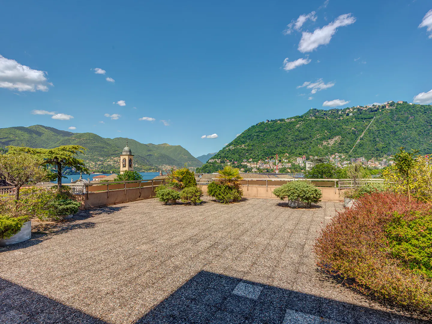Rooftop view of Lake Como, Italy, with mountains, blue sky, and a church tower in the background. The rooftop is paved and has shrubs.
