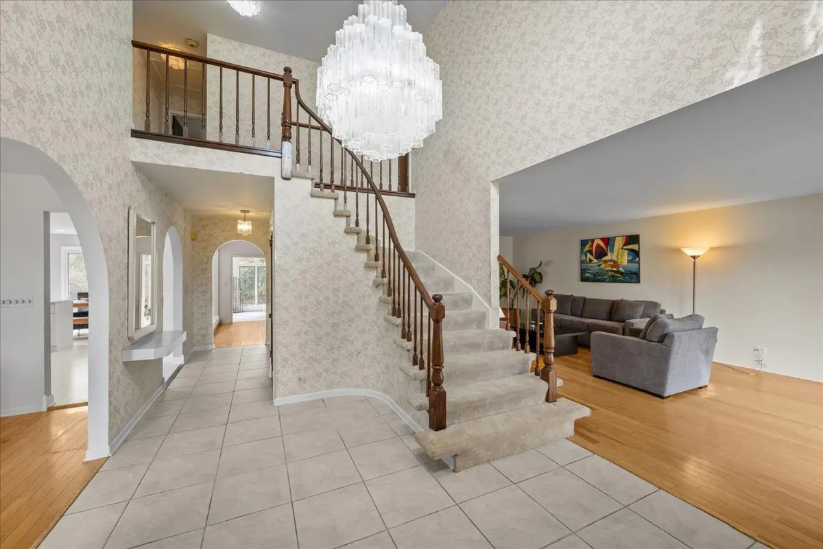Foyer view with a staircase, chandelier, and arched doorways. Living room with gray sofas and a sailboat painting is visible.